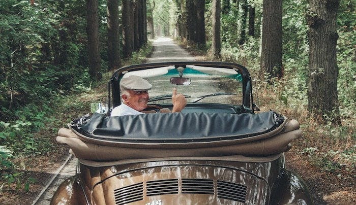 An elderly man sits in a vintage car with its roof down. He's looking back at the camera with a thumb up and a smile.