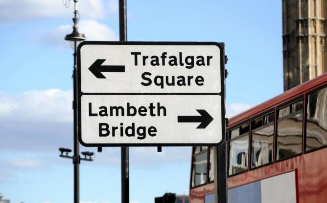 Photograph of a directional road sign in London. There is arrow pointing left to Trafalgar Square, and another arrow pointing right to Lambeth Bridge.