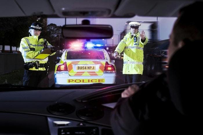 A photograph taken at night from a car's interior facing out through the windshield. Two police officers stand in front of the car, with the police car behind in the background. The police car's lights are flashing and an officer is holding up his hand palm-out to indicate the driver must stop.