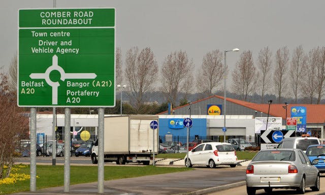 Photograph of cars approaching a roundabout, with a road sign in the foreground. The road sign reads 'COMBER ROAD ROUNDABOUT'. The first exit is to Belfast. The second exit is to the Town Centre and Driver and Vehicle Agency. The third exit is to Bangor and Portaferry.