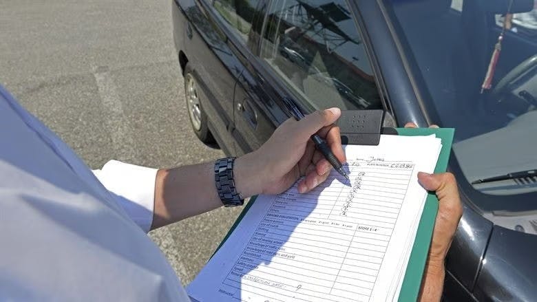 A driving test examiner stands next to a car and makes notes on a driving test mark sheet 